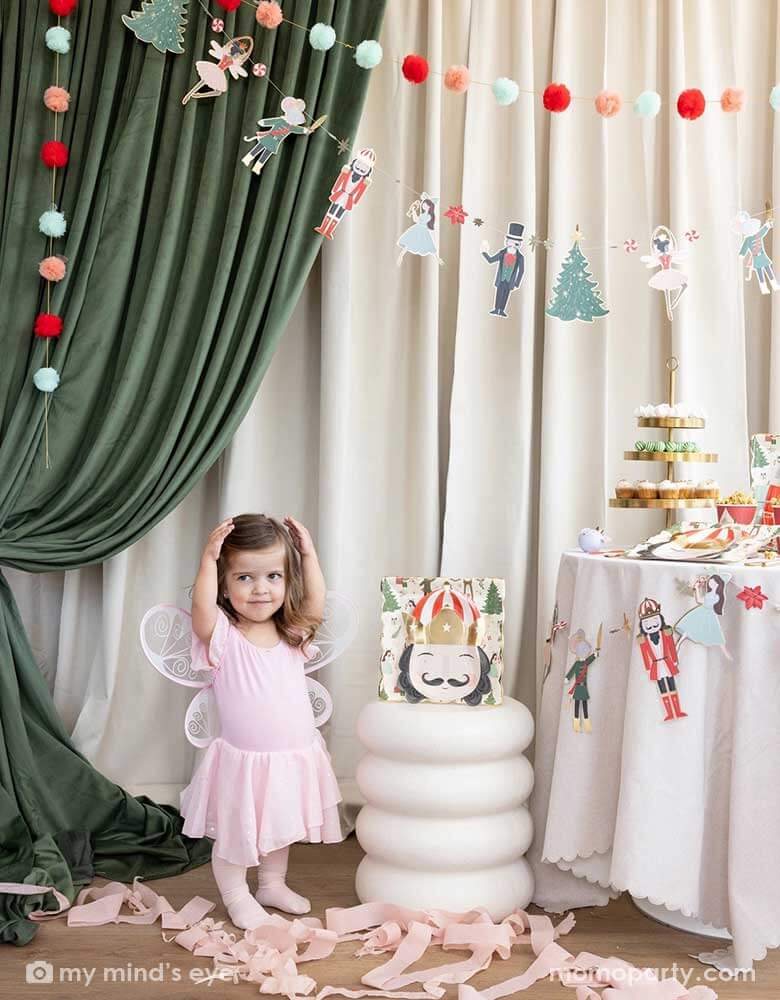 A little girl doing a ballet pose standing next to a Holiday party table filled with Holiday treats and Nutcracker themed party supplies including party plates, cups, party garland. In her back there's a stage curtain in satin green and beige which is decorated with Momo Party's Nutcracker garland with pom-poms which adds a festive touch to the Holiday decoration.