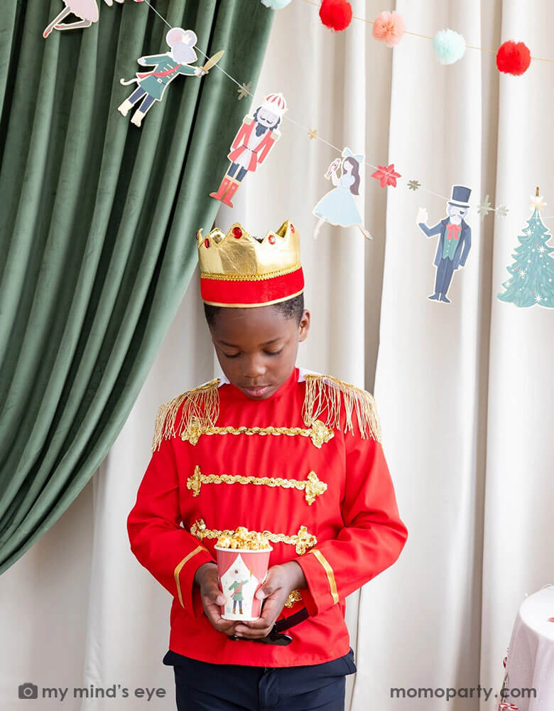 A boy dressed up as Nutcracker's soldier holding Momo Party's 12 oz Nutcracker party cup filled with popcorn. In his back there's a stage curtain in satin green and beige which is decorated with Momo Party's Nutcracker garland with pom-poms which adds a festive touch to the Holiday decoration.