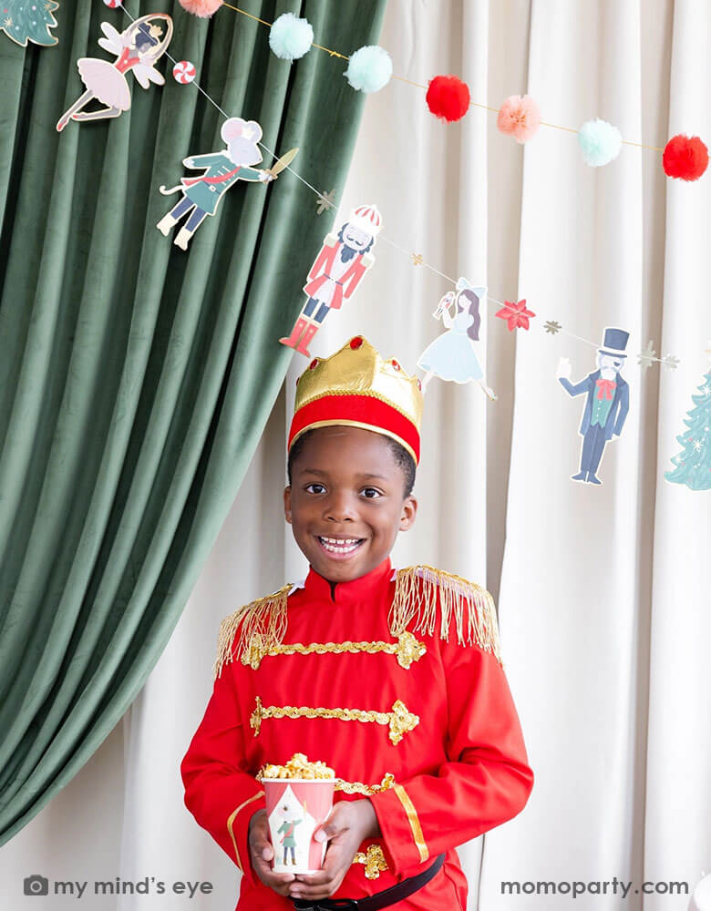 A boy dressed up as Nutcracker's soldier holding Momo Party's 12 oz Nutcracker party cup filled with popcorn. In his back there's a stage curtain in satin green and beige which is decorated with Momo Party's Nutcracker garland with pom-poms which adds a festive touch to the Holiday decoration.