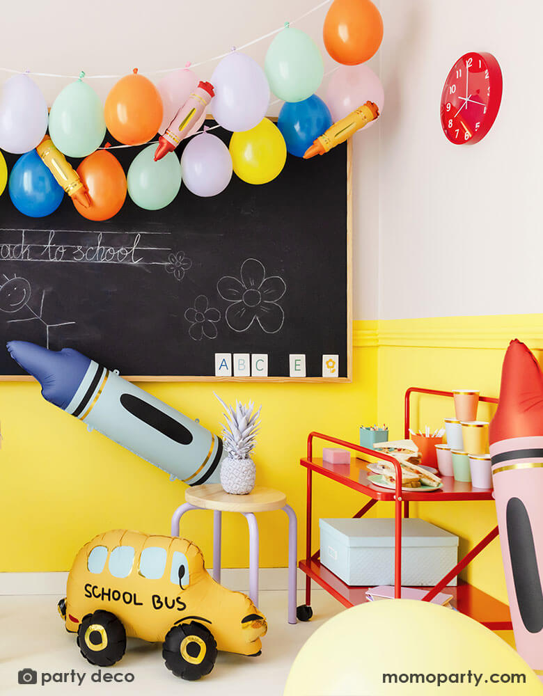 A school classroom set up decorated with festive party balloons in various colors on the chalkboard. The balloon garland is decorated with mini crayon shaped balloons in multiple colors. Around the the chalkboard are some large sized crayon shaped foil balloons in blue and red and a school bus shaped foil balloon on the floor. Along with colorful vibrate school furnitures, making this a perfect decorating inspo for a classroom decoration to welcome the new school year!