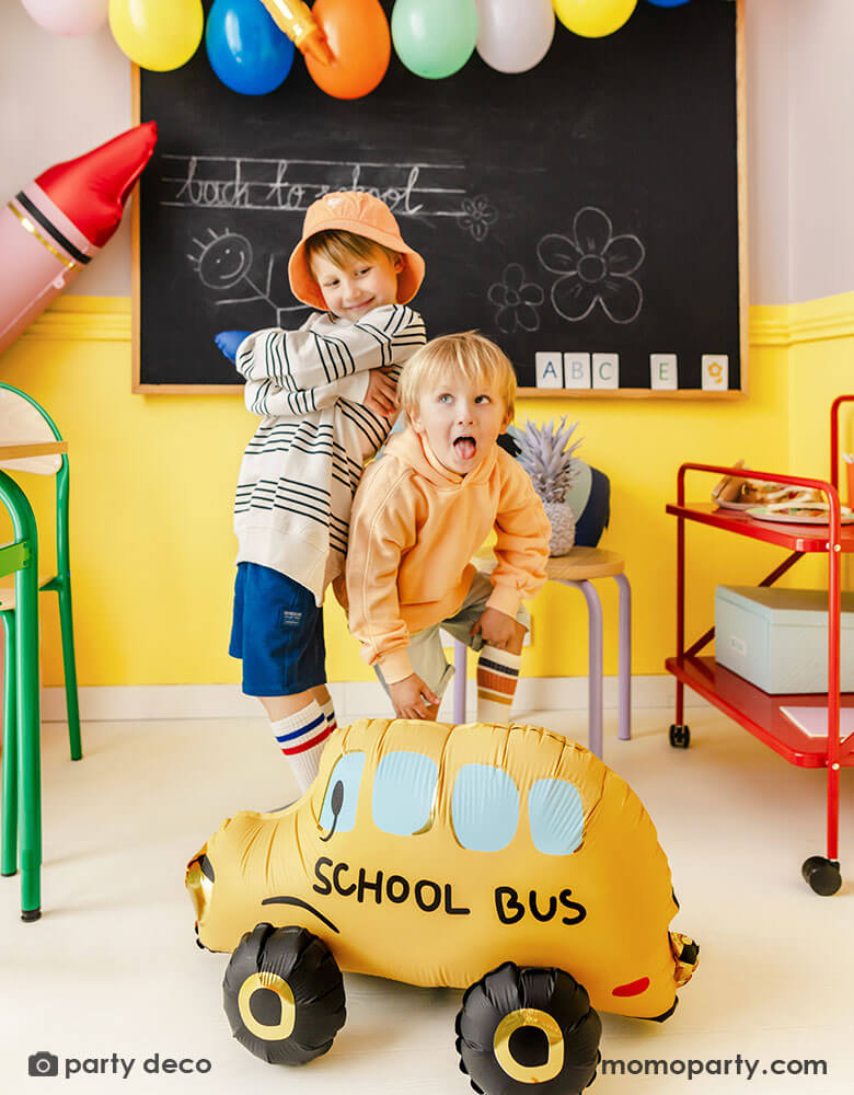 Two school aged boys making funny faces in a school classroom setting decorated with Momo Party's back to school party balloons including a school bus shaped foil balloon, a red crayon shaped foil balloon, a few festive colorful latex balloons hung above the chalkboard. With colorful, vibrant school furniture around it, this makes a great inspo for a modern and playful back to school, first day of school celebration in a school set up.