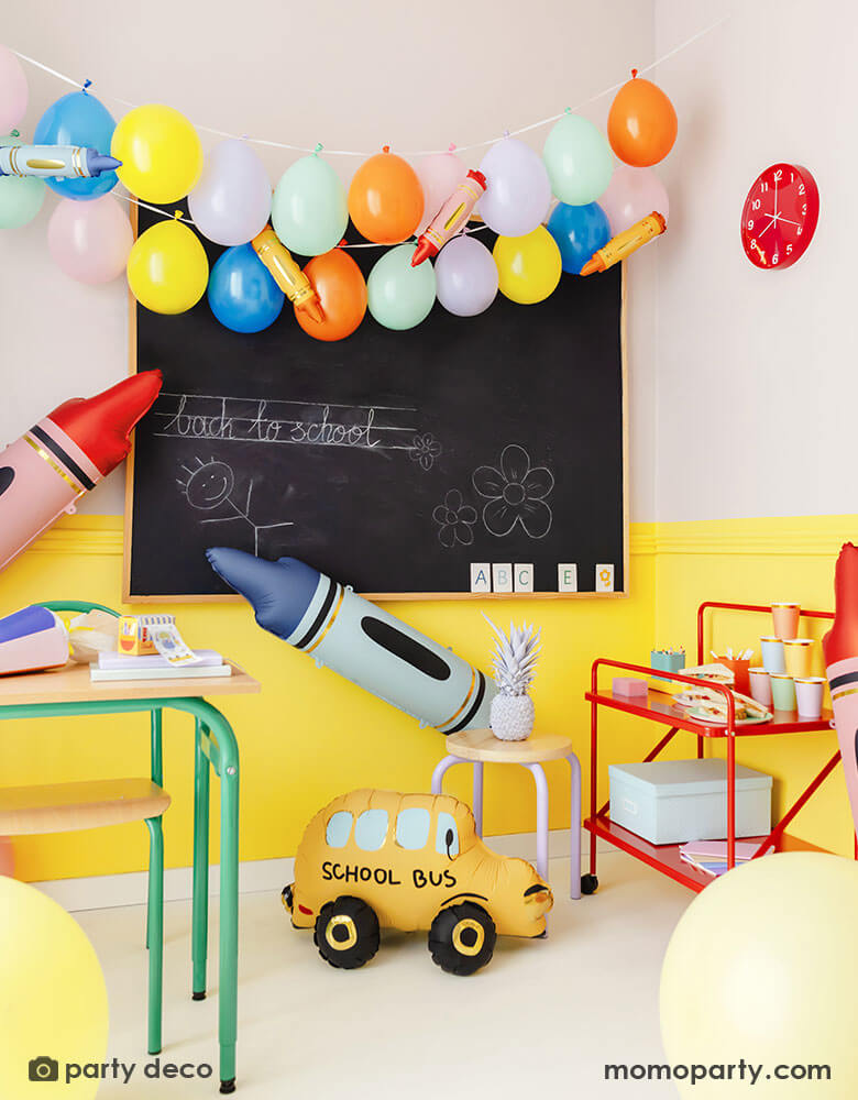 A school classroom set up decorated with festive party balloons in various colors on the chalkboard. The balloon garland is decorated with mini crayon shaped balloons in multiple colors. Around the the chalkboard are some large sized crayon shaped foil balloons in blue and red and a school bus shaped foil balloon on the floor. Along with colorful vibrate school furnitures, making this a perfect decorating inspo for a classroom decoration to welcome the new school year!