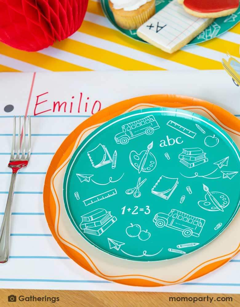 A festive back to school first day of school celebration party table place setting featuring Momo Party's back to school themed party supplies including the school day assorted side plates with school supplies designs in green, and the loose leaf paper placemat by Gatherings. With school supplies shaped sugar cookies around and a honeycomb apple decoration on the table, makes a great inspo to celebrate the start of the new school year with kids!