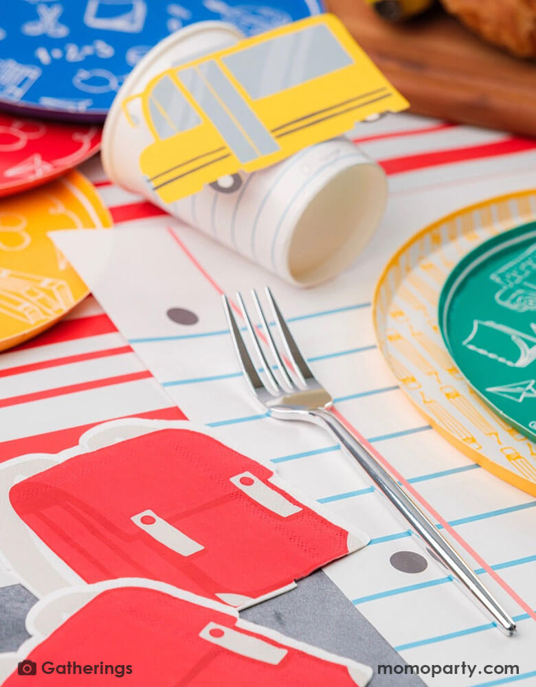 A festive back to school first day of school celebration party table setting featuring Momo Party's back to school themed party supplies including the No. 2 pencil patterned dinner plates, school day side plates with school supplies designs in four different colors, and the notebook inspired placemat by Gatherings. With pencil shaped napkins and red backpack shaped napkins, plus a plate of treats and fruits, this makes a great inspo to celebrate the start of the new school year with kids!