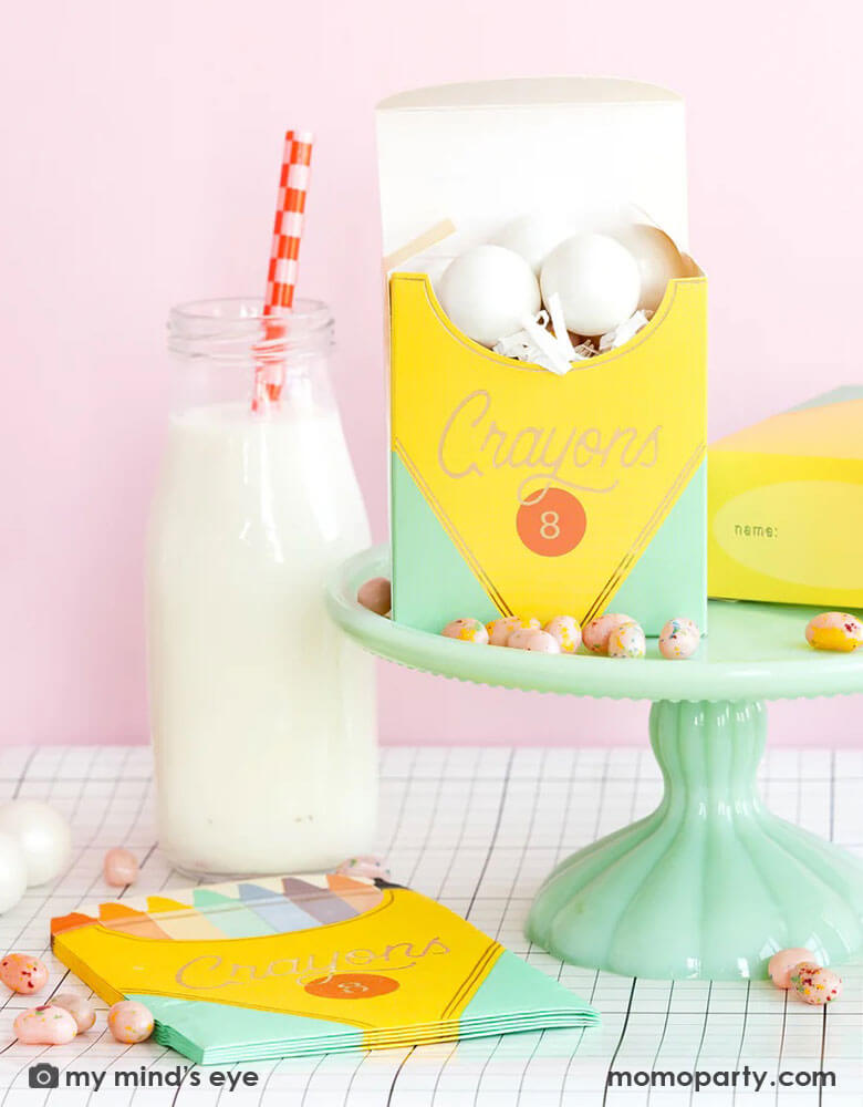 A back to school party table set up in a pink room featuring school themed party supplies from Momo Party including crayon box shaped napkins, crayon box treat boxes by My Mind's Eye on a mint cake stand on a grid table runner next to a milk bottle, with jelly beans around, this makes a great inspiration for a first day of school celebration or kid's back to school parties.