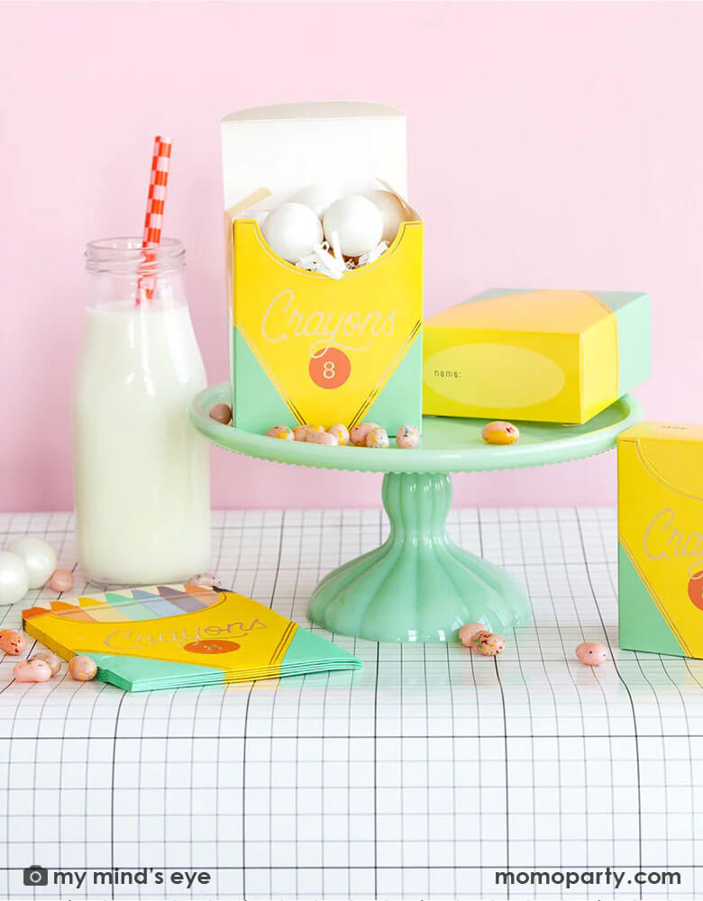 A back to school party table set up in a pink room featuring school themed party supplies from Momo Party including crayon box shaped napkins, crayon box treat boxes by My Mind's Eye on a mint cake stand on a grid table runner next to a milk bottle, with jelly beans around, this makes a great inspiration for a first day of school celebration or kid's back to school parties.