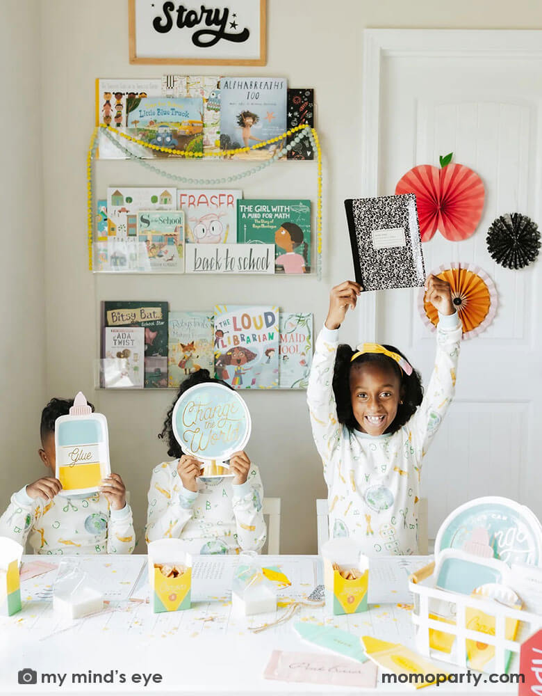 A first day of school breakfast party with three siblings in their school themed pajamas at their house. The kids are holding the notebook shaped plate, the globe shaped plate, the glue bottle shaped plate. On the table there are more school themed party supplies from Momo Party including crayon shaped treat boxes, eraser shaped napkins, notebook shaped plates. On the wall there are also some apple and pencil shaped party fans as the backdrop. A simple yet cute way to celebrate learning!