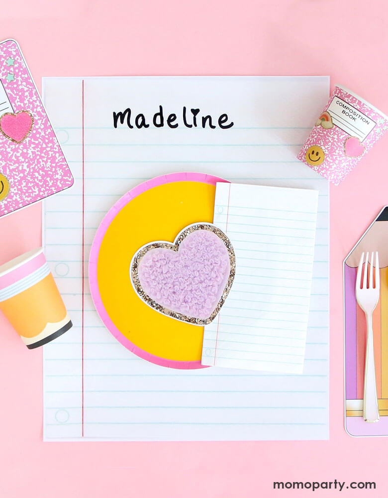 An adorable pink party table features Momo Party's back to school party supplies by Kailo Chic including the huge notebook placemat with the name "Madeline" written on it. notebook shaped napkins, pink composition book party cups, pencil paper cups and a round pink and yellow block plates inspired by pencils, making this a perfect inspo for kid's back to school party or first day of school celebration.