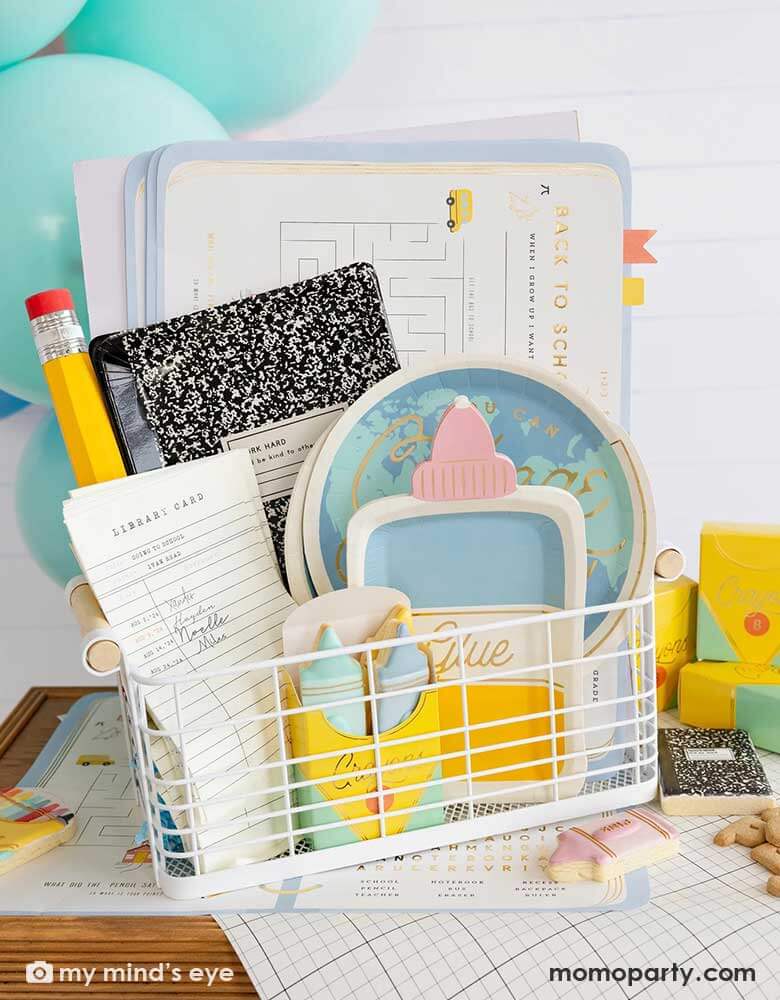 A back to school occasions bin filled with school themed party supplies from Momo Party including back to school planner placemats, globe shaped plates, glue bottle shaped plates, library card shaped napkins, a crayon box shaped treat box filled with crayon shaped sugar cookies and a giant pencil. On the table there's a notebook grid inspired table runner. With the party balloons in the back, it's a great inspo for a festive kid's first day of school celebration party!