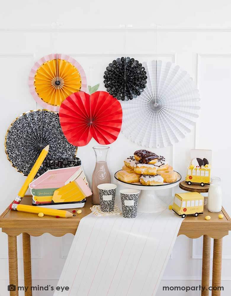 A school themed party table featuring school themed party supplies from Momo Party including stack of books plates, pencil shaped napkins, composition notebook party cups, school bus treat boxes and writing paper table runner around some donuts and milk bottles. The back wall is adorned with back to school party fans by My Mind's Eye including an apple, an pencil, a notebook, an equation pattern and a composition notebook fan. A perfect inspo for kid's back to school, first day of school celebration.