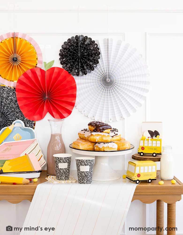 A party table featuring school themed party supplies from Momo Party including stack of books plates, backpack plates, pencil shaped napkins, composition notebook party cups, school bus treat boxes and writing paper table runner around some donuts and milk bottles. The back wall is adorned with back to school party fans by My Mind's Eye including an apple, an pencil, a notebook, an equation pattern and a composition notebook fan. A perfect inspo for kid's back to school, first day of school celebration.