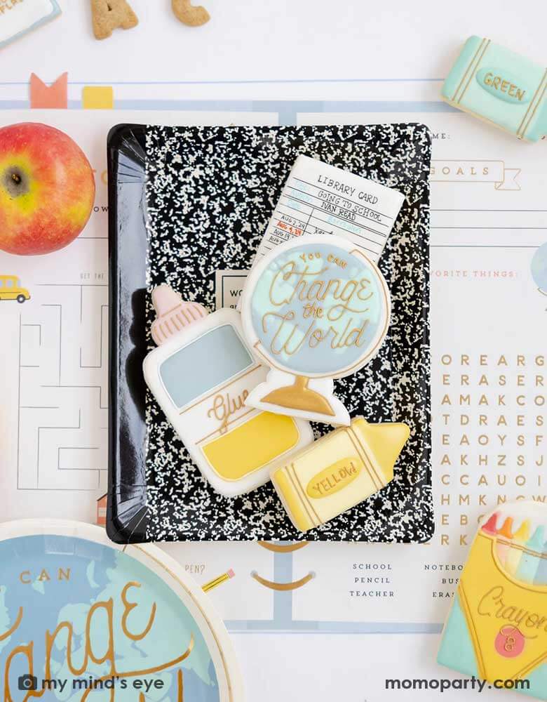 A overhead shot of a place setting from a back to school party featuring Momo Party's school themed party supplies including the back to school planner placemats, a black notebook shaped plate which has multiple matching school supplies themed sugar cookies including crayons, a glue bottle, a globe, a library card on it, with an apple and some alphabet shaped cookies over the placemat, this makes a festive and adorable table setting ideas for kid's first day of school celebration.