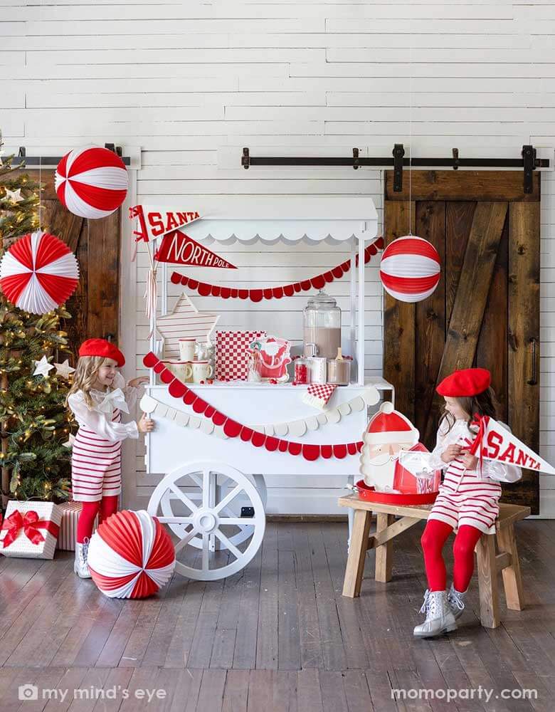 Two girls in an adorable Santa themed hot cocoa station set up for a kid's Holiday party featuring a white dessert cart with hot chocolate and Momo Party's Dear Santa party supplies including Santa shaped party cups, checkered plates, napkins, Santa on a sleigh shaped plates and Dear Santa envelope shaped plates. Around the hot chocolate station are two felt party pennant with Santa & Northpole written on them, red scalloped shaped party garlands and some red and white honeycomb balls hung from the ceiling.