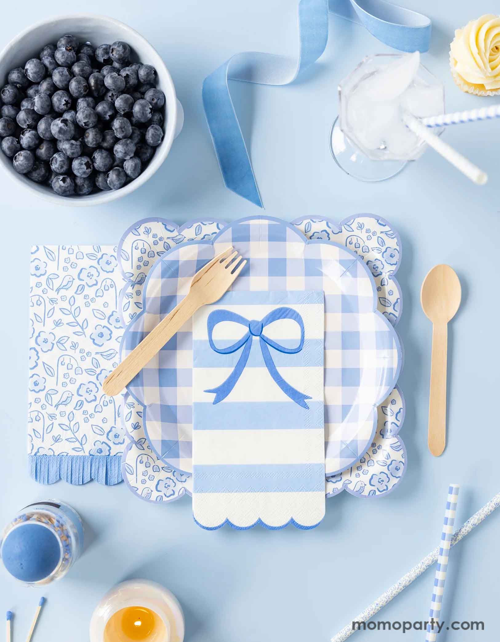 A blue inspired tablescape featuring Momo Party's blue party tableware collection by My Mind's Eye. Featuring various party plates, napkins and party straws in different sizes, shapes and spring inspired patterns including gingham checkered, elegant bows, and floral toile. Along with blue ribbons, blueberries and some cupcakes on the table, this tablescape is a perfect inspiration for your spring gatherings or Easter celebrations!