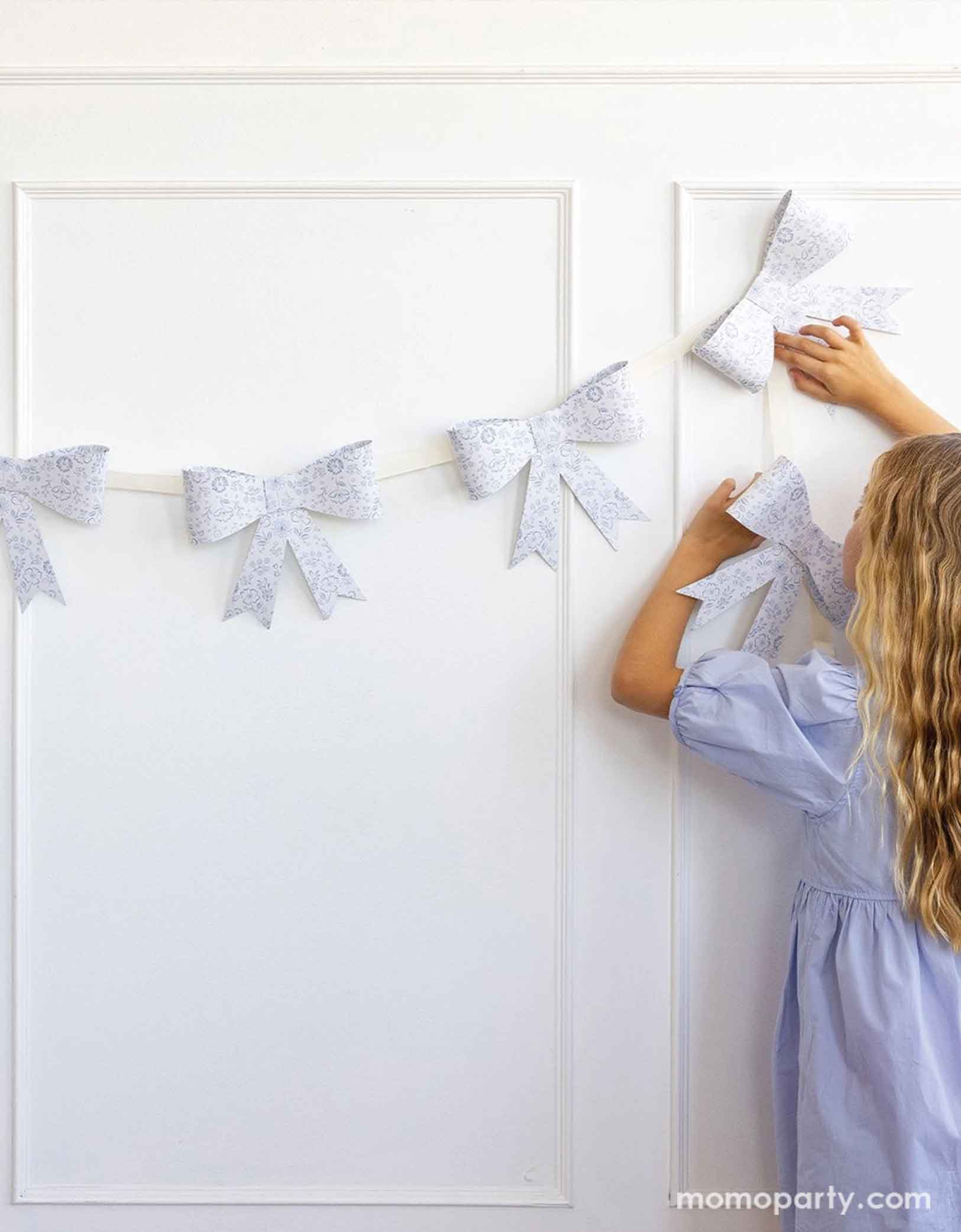 A girl in pastel blue dress holding and hanging Momo Party's 7' Floral Bow Banner onto a white wall. Made of high-quality paper, this garland features delicate flowers and a cute bow design. Perfect for weddings, baby showers, or garden parties. Spring into style with this beautiful banner!