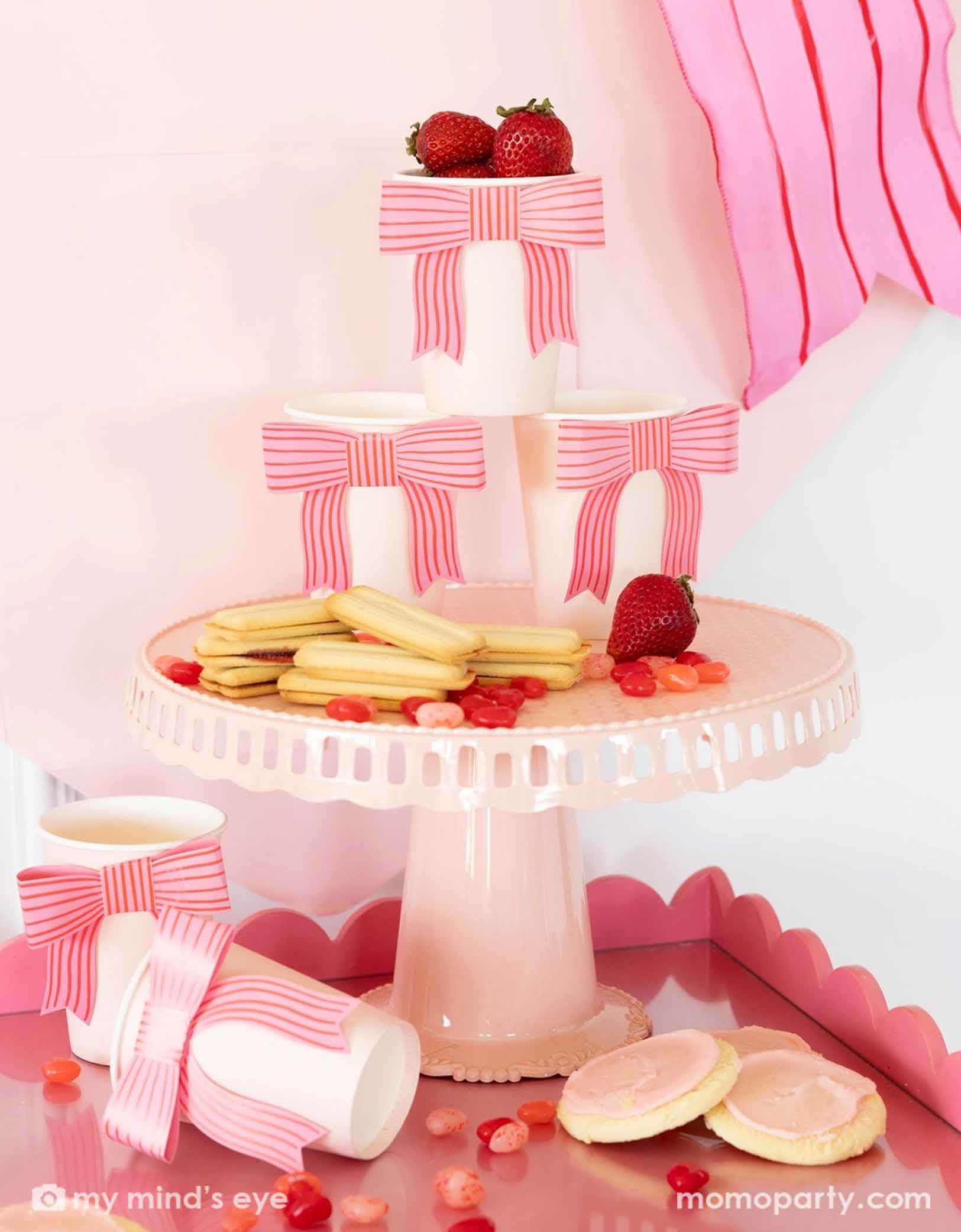 A gorgeous Valentine's Day or Galentine's Day party set up featuring Momo Party's bow party cups staggered on a pink cake stand. On the tray and the stand there are pink and red themed desserts / snacks including sugar cookies, strawberries, jelly beans and more. Making this a great inspiration for a darling Vday celebration set up!