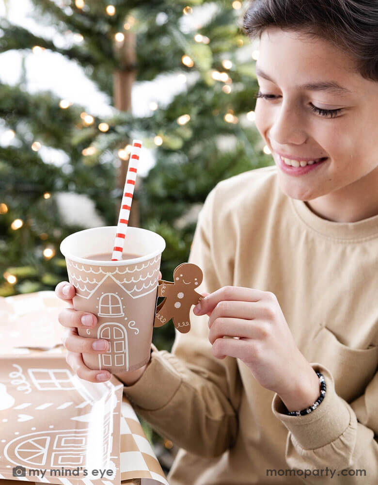 A boy is holding and looking into Momo Party's 12 oz Gingerbread Handled Paper Cup with a festive gingerbread man handle. There's hot chocolate in the cup with a red and white striped reusable straw. With gingerbread man shaped plates and a Christmas tree with lights in background, this creates a cozy yet festive vibe for a whimsical Holiday party for kids.