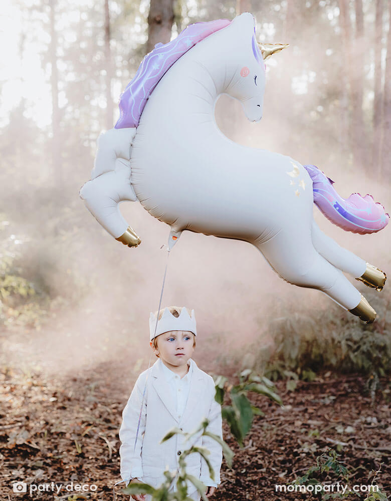 A boy dressed in white clothing with a party crown hat in an enchanting forest with Momo Party's enchanting unicorn shaped foil balloon floating in the air, making this a dreamy scene for kid's special celebration.