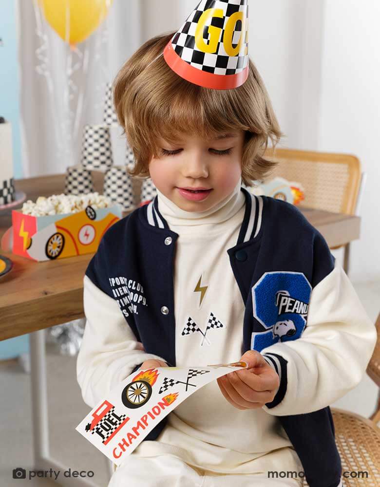 A little boy wearing a checkered flagged party hat with GO! on it in a race car party. In his back is a party table filled with race car themed treat boxes with popcorn in them. He's holding Momo Party's race car sticker sheets by Party Deco - a perfect party activity for boy's race car themed birthday celebrations.