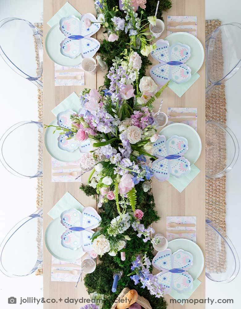 An overhead shot of a beautiful butterfly themed party table featuring Momo Party's Flutter butterfly shaped plates paired with pastel mint dinner plates and napkins, next to it is butterfly themed party cups and napkins. In the middle there's moss grass textured table runner with pretty spring flower arrangement. On the wall there are butterfly shaped paper decorations attached on the wall, making this a perfect inspo for kid's butterfly fairy themed birthday or a gorgeous spring Easter celebration. 