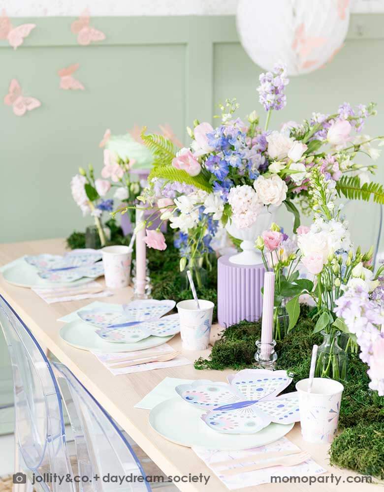 A beautiful butterfly themed party table featuring Momo Party's Flutter butterfly shaped plates paired with pastel mint dinner plates and napkins, next to it is butterfly themed party cups and napkins. In the middle there's moss grass textured table runner with pretty spring flower arrangement. On the wall there are butterfly shaped paper decorations attached on the wall, making this a perfect inspo for kid's butterfly fairy themed birthday or a gorgeous spring Easter celebration. 