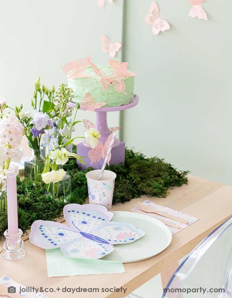 A beautiful butterfly themed party table featuring Momo Party's Flutter butterfly shaped plates paired with pastel mint dinner plates and napkins, next to it is butterfly themed party cups and napkins. In the middle there's moss grass textured table runner with pretty spring flower arrangement and paste pink candles. On the wall there are butterfly shaped paper decorations attached on the wall, making this a perfect inspo for kid's butterfly fairy themed birthday or a gorgeous spring Easter celebration. 