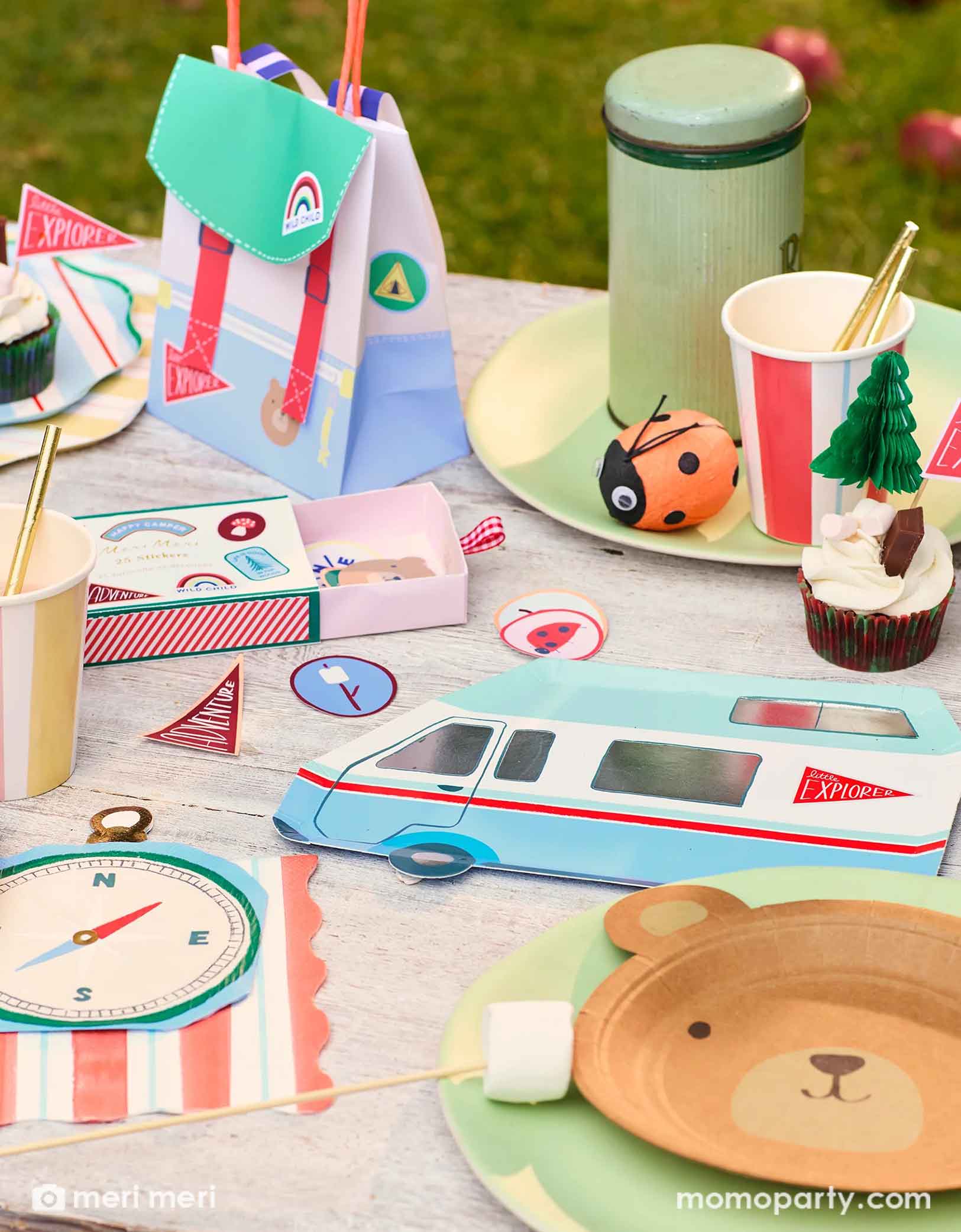 A camping themed birthday party table featuring Momo Party's camping themed party supplies by Meri Meri including the camping RV van shaped plate, the camping compass shaped napkins, the camping themed stickers, and camping backpack shaped party bag, along with striped dinner plates, party cups and a vintage thermos bottle with a ladybug surprise ball next to it, this camping adventure party collection is perfect for your nature-loving child's camping themed celebration.