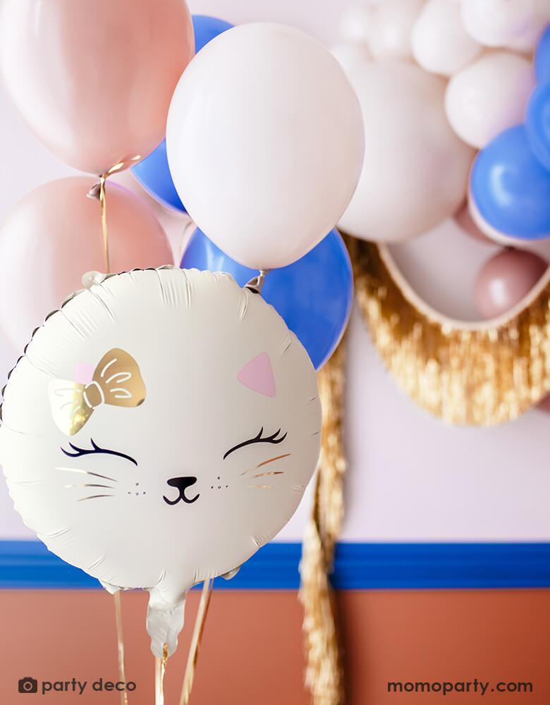 A cat themed party decoration set up features Momo Party's 18" kitty cat shaped foil balloon in cream, along with some latex balloons in pink, blush and royal blue adorned with gold tinsel garland hung on the wall, making this an adorable inspo for kid's kitty cat or pet themed party decoration.