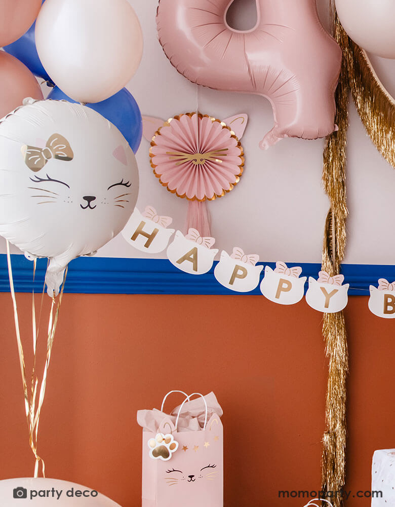 A kid's cat themed birthday party decoration set up features Momo Party's cat themed party supplies by Party Deco including 18" kitty cat shaped foil balloon in cream, along with some latex balloon mix in pink, blush and royal blue. On the wall there are pink kitty cat shaped party fans along with a gold tinsel garland hung on. With a pink number 4 foil balloon and a cat head shaped happy birthday banner, it makes an adorable inspo a cat or pet themed party.