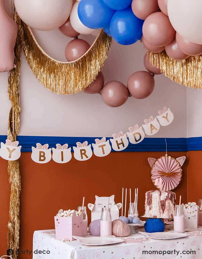 A kid's kitty cat themed party decorated with pink cat shaped paper fans, gold tinsel garland, pink and blue balloons and pink happy birthday banner. In the front there's a table filled with cat themed party supplies including cat shaped plates, treat boxes and pink paw shaped napkins.