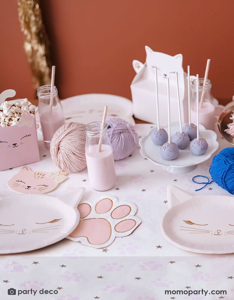 A cat themed party table featuring various cat themed party supplies from Momo Party including party plates, pink paw shaped napkins, cat shaped treat boxes filled with popcorn, small milk bottles with pale pink drink and a table of treats and snacks in pastel pink and lilac colors, making this an adorable inspo for kid's kitty cat themed birthday celebration.