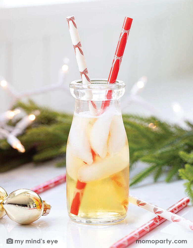 A close up of a glass filled with apple juie with Momo Party's red and white Christmas Bows reusable straws by My Mind's Eye. Around the table there's a Christmas wreath and some Christmas tree ornaments in gold. Making this a festive vibe for the Holiday season. 