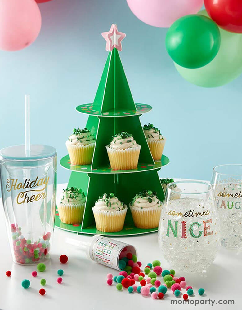 A festive Holiday party table features Momo Party's Christmas tree shaped cupcake stand with cupcakes with green sprinkles on them. On the table there are some tumblers and cups and a confetti cannon filled with mini pom poms in Christmas themed color, i.e red, green, and pink. Behind the table are some festive party balloons in red, pink, green and mint making this a perfect inspo for a festive Christmas party table ideas.