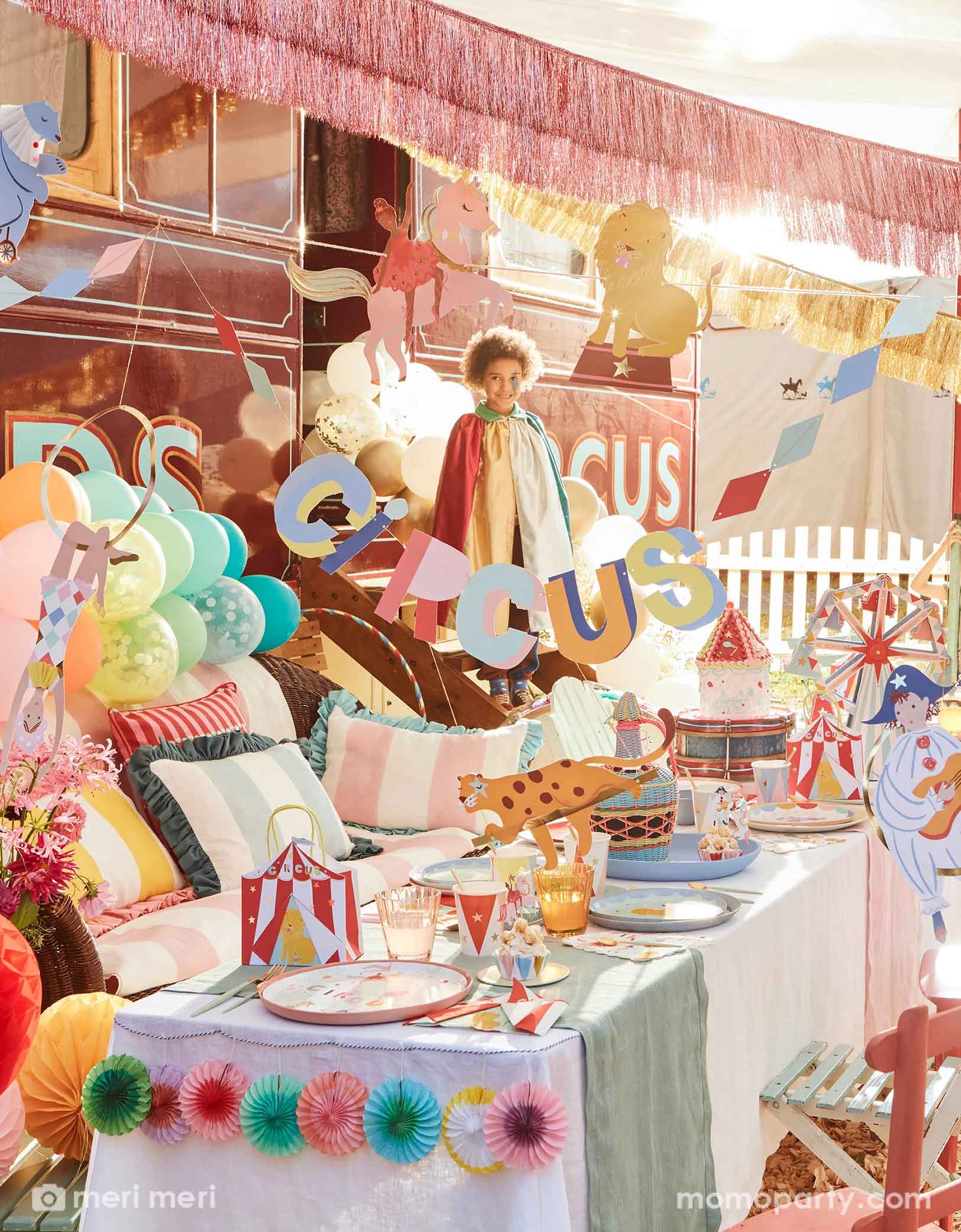 A festive circus themed party featuring Momo Party's circus themed party supplies including a table filled with circus themed party tablewares including plates, napkins, party cups, party bags and ferry's wheel cupcake stand. There's the circus party garland featuring circus animals with honeycomb decorations hung above the party table. Above the party garland, there are pink and gold fringe garlands. Around the table there are some kids dressed up in circus inspired costumes, making this a playful scene.  