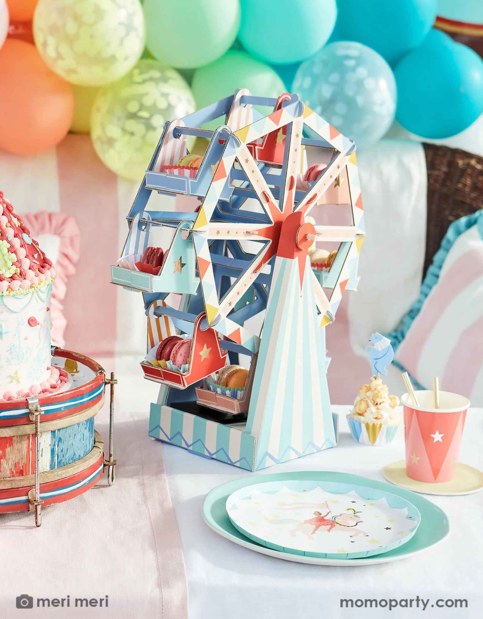 A festive carnival, circus themed party featuring Momo Party's circus themed party supplies and decorations by Meri Meri including the circus side plates, party cups, cupcake kit, ferry's wheel cupcake stand and a rainbow colored balloon garland in the back, with a circus tent shaped birthday cake on a vintage drum and pastel striped table cloth, this makes a great inspiration for a playful and fun circus party decoration idea.