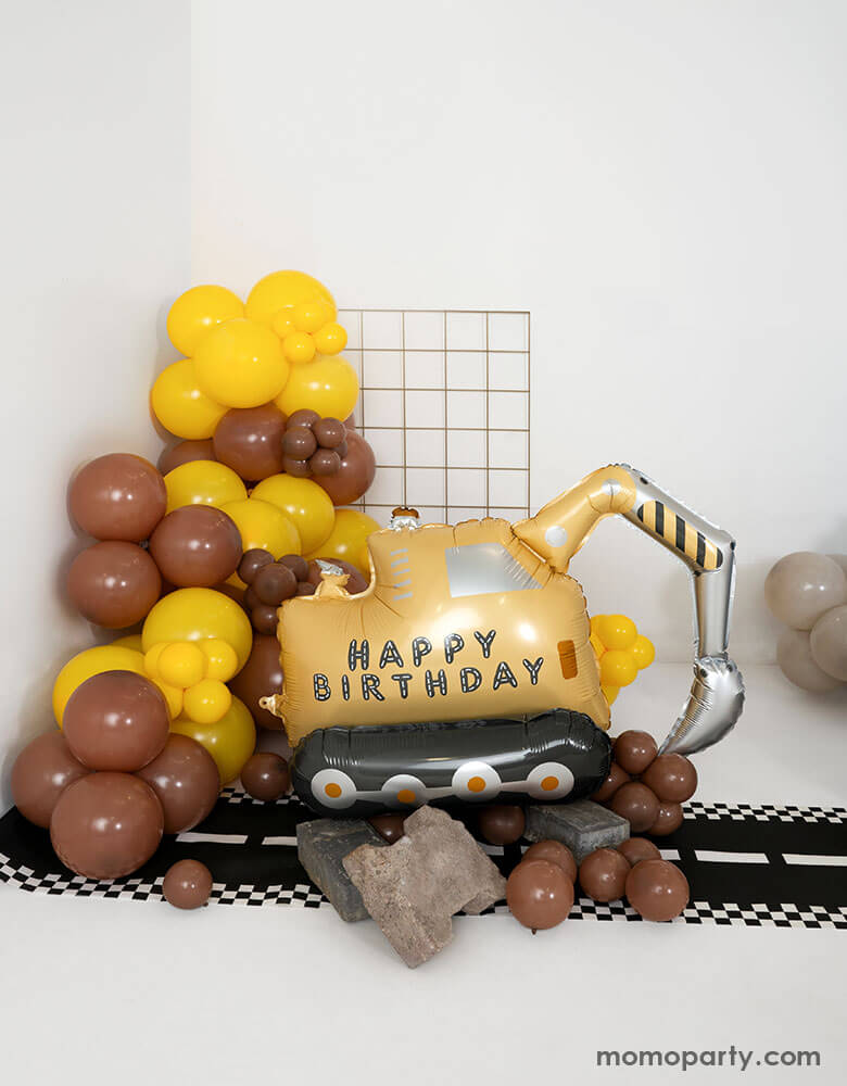 A construction themed birthday party balloon set up featuring Momo Party's digger shaped happy birthday foil balloon next to a balloon garland in brown and yellow. On the floor there's a road table runner. Making this a fun decoration for kid's construction themed birthday celebration!