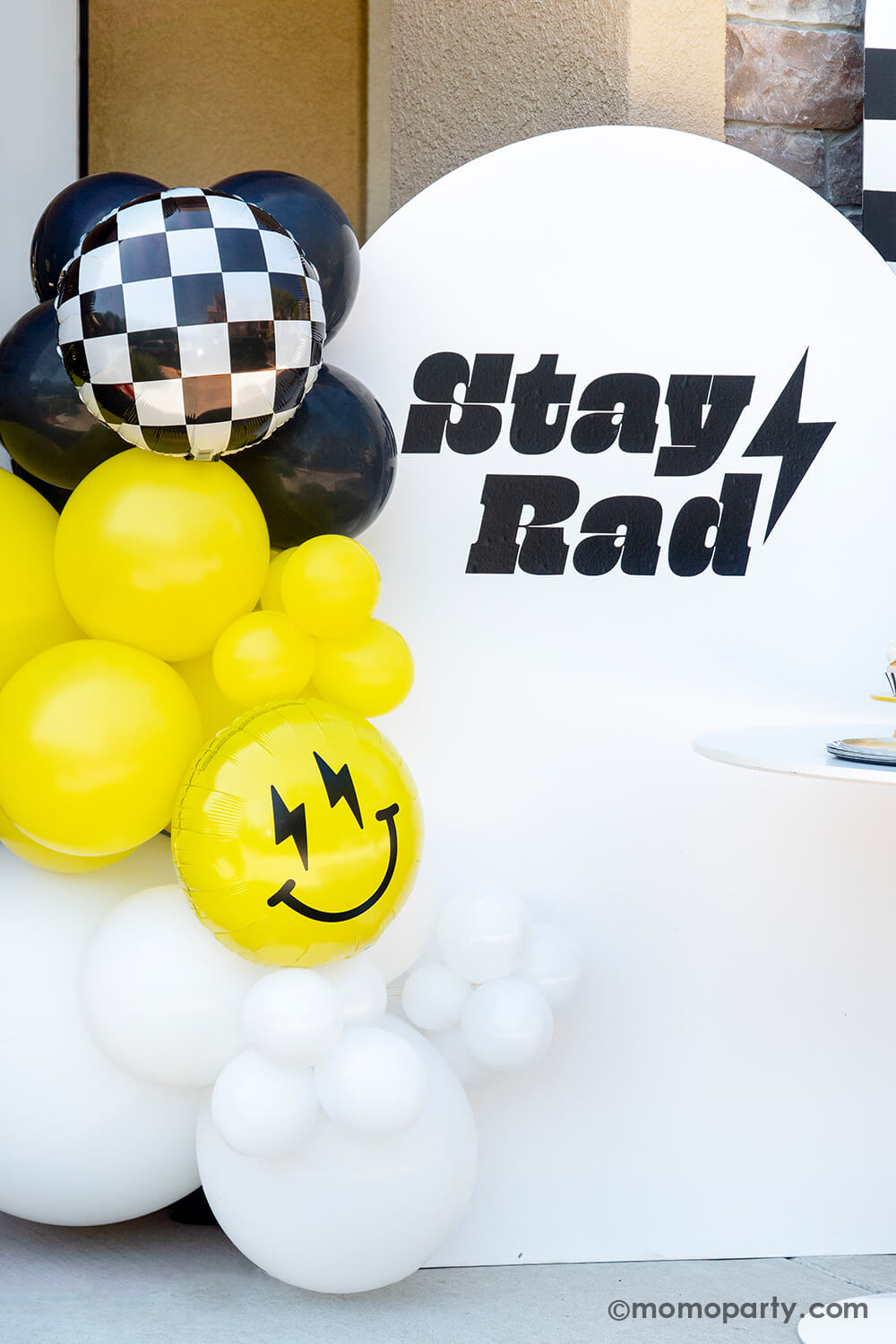 A close up look of Momo Party's "Cool Dude" themed Birthday Party, featuring a balloon garlands in black, white and yellow adorned with a black checkered board foil balloon and a smiley fact foil balloon with lightning bolt eyes, in front of white wooden backdrop panel adorned with "Stay Rad" and a lightning bolt sticker. This decoration is perfect for boy's "One Happy Dude" themed first birthday, a "Two Cool" themed second birthday party or "Ten Rad Years" 10th birthday party.