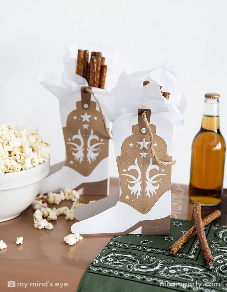 A cowboy themed party table filled with treats and snacks and Momo Party's cowboy boot shaped treat boxes filled with pretzel sticks. On the table there's a soda bottle and a green bandana as decoration, perfect for a Western rodeo themed celebration, be a First Rodeo boy's first birthday party or a fun cowboy bash!