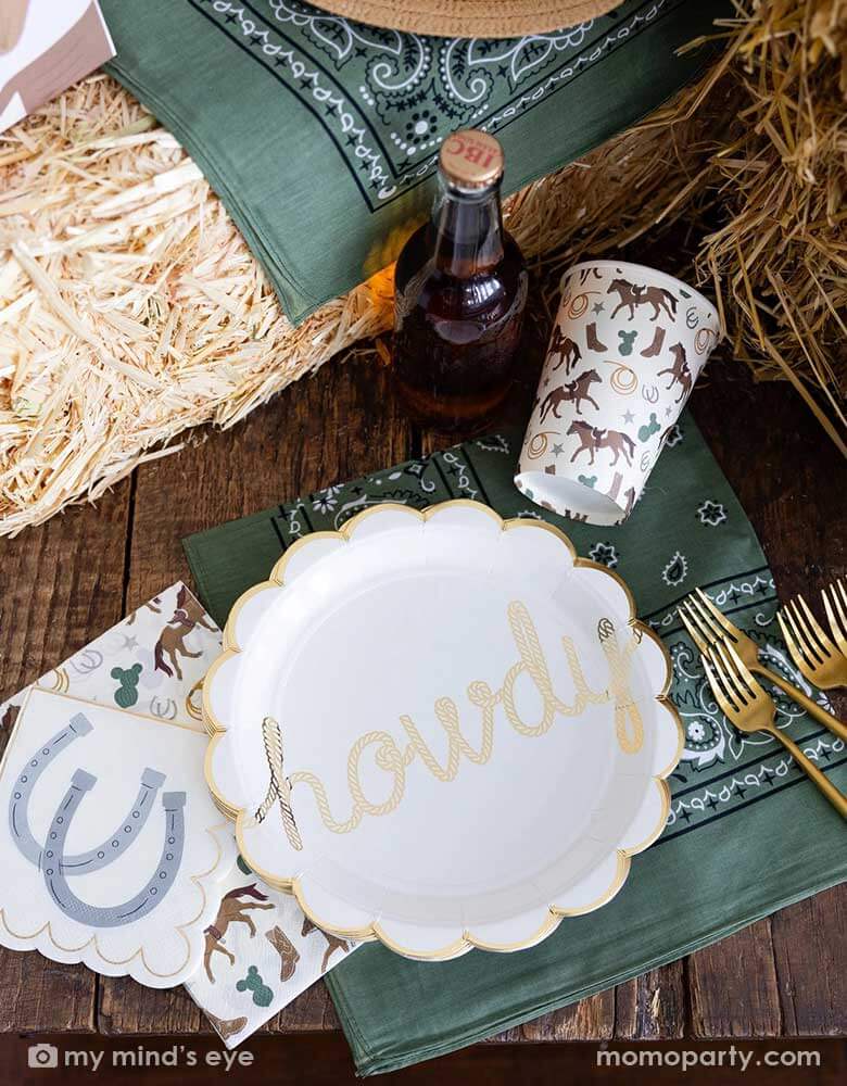 An overhead shot of a cowboy themed party tablescape featuring Momo Party's cowboy themed party supplies including howdy paper plates, cowboy patterned cups, and napkins. On the table there's a straw bale, and a green bandana as decoration, and a soda bottle, perfect for a Western rodeo themed celebration, be a First Rodeo boy's first birthday party or a fun cowboy bash!