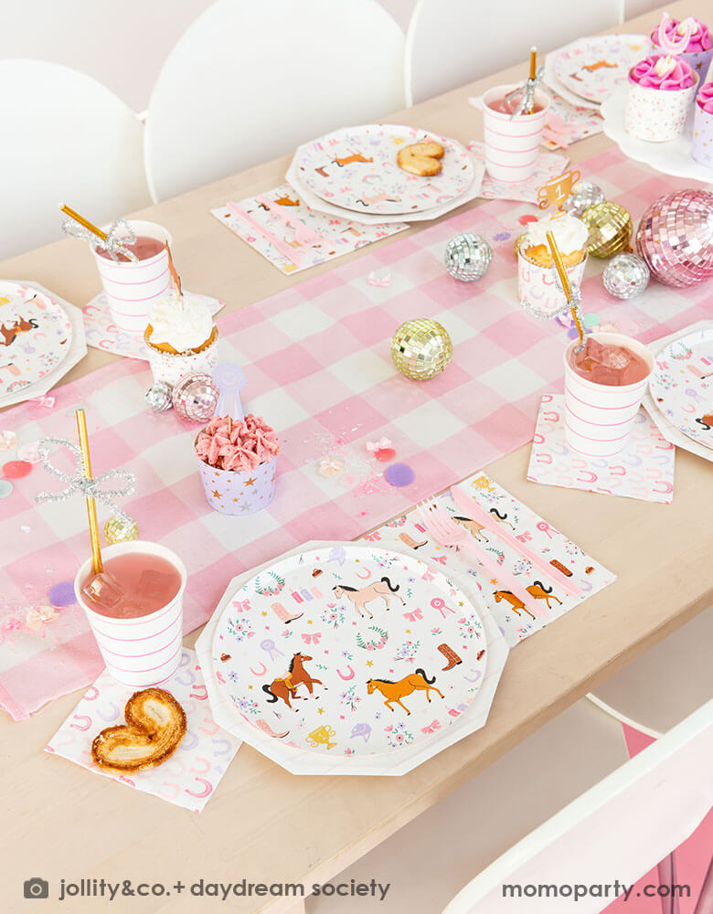 A girly cowgirl rodeo pony themed party table featuring Momo Party's cowgirl pony themed party plates, napkins, horseshoe napkins, pink striped party cups and a pink gingham table runner. Around the tableware there are disco ball decorations and a cake stand with cupcakes with pink icing, making this an adorable inspiration for girl's First Rodeo first birthday celebration or an adorable cowgirl theme party for pony-loving girls!