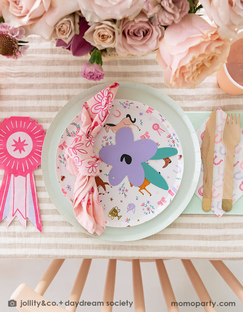 An overhead shot of a girly pony themed party tablescape featuring Momo Party's cowgirl pony themed party plate, horseshoe napkin, pink ribbon award shaped napkin and gold dipped wooden cutlery, with a pink bandana, all on a mocha brown striped tablecloth. There's some beautiful rose bouquet as the centerpiece, making this an adorable inspiration for girl's pony horse themed birthday celebration.