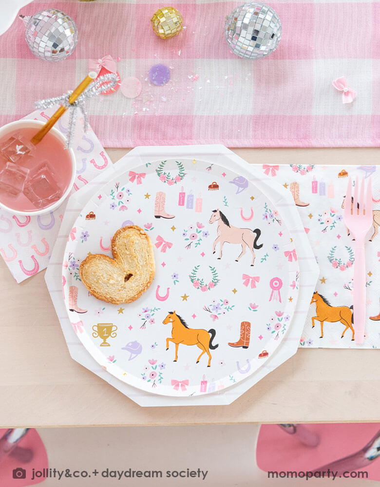 A close up of a girly cowgirl rodeo Western themed party tablescape featuring Momo Party's cowgirl pony themed party plate, napkin, horseshoe napkin, pink striped party cups and a pink gingham table runner. Around the tableware there are some lilac round confetti spread-out and a small pink disco ball decoration, making this an adorable inspiration for girl's First Rodeo first birthday celebration or an adorable cowgirl theme party for pony-loving girls!