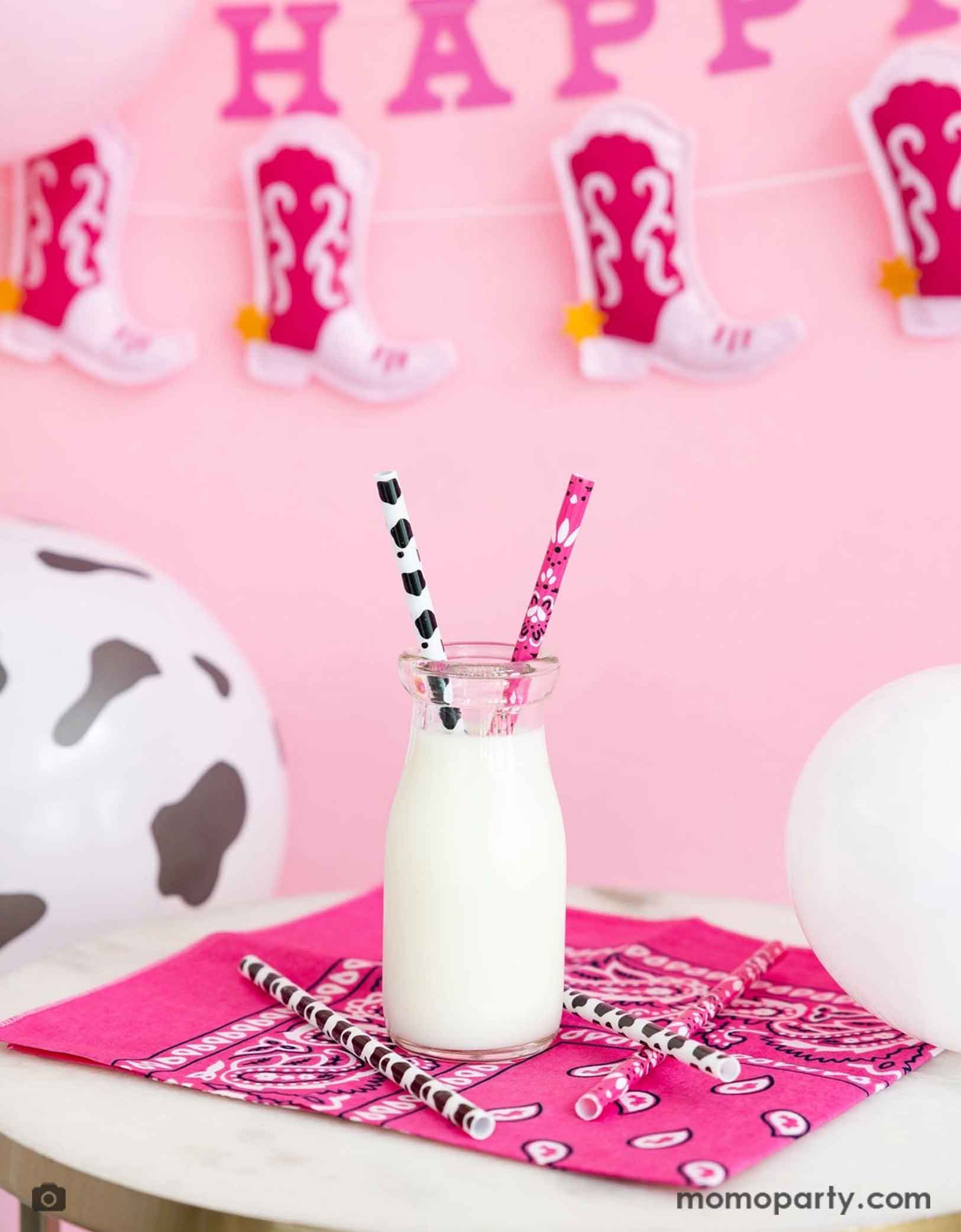 A fun cowgirl party table featuring a hot pink bandana with a bottle of milk with Momo Party's cowgirl party reusable straws in pink and cow print design. In the back of the party table are some cow print balloons and a cowgirl boot Happy birthday party banner hung on a pink wall. Making this a fun yet simple decorating idea for a cowgirl disco celebration - whether it's a "First Rodeo" girl's first birthday party or a fun cowgirl bachelorette party.