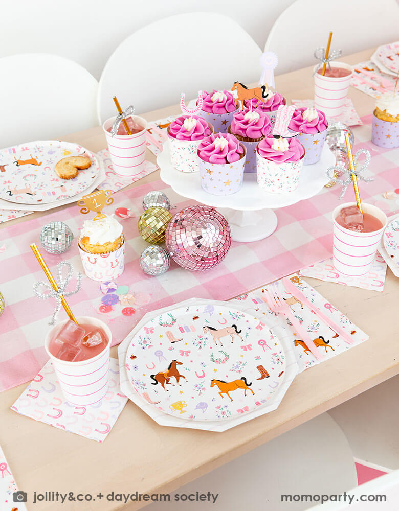 A girly cowgirl rodeo pony themed party table featuring Momo Party's cowgirl pony themed party plates, napkins, horseshoe napkins, pink striped party cups and a pink gingham table runner. Around the tableware there are disco ball decorations and a cake stand with cupcakes with pink icing, making this an adorable inspiration for girl's First Rodeo first birthday celebration or an adorable cowgirl theme party for pony-loving girls!