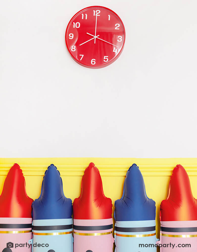 A line up of Momo Party's blue crayon shaped foil balloon and red crayon shaped foil balloon by Party Deco against a yellow wall. On the wall there's a red school clock at 8 oclock, creating a first day of school scene for kid's back to school season.