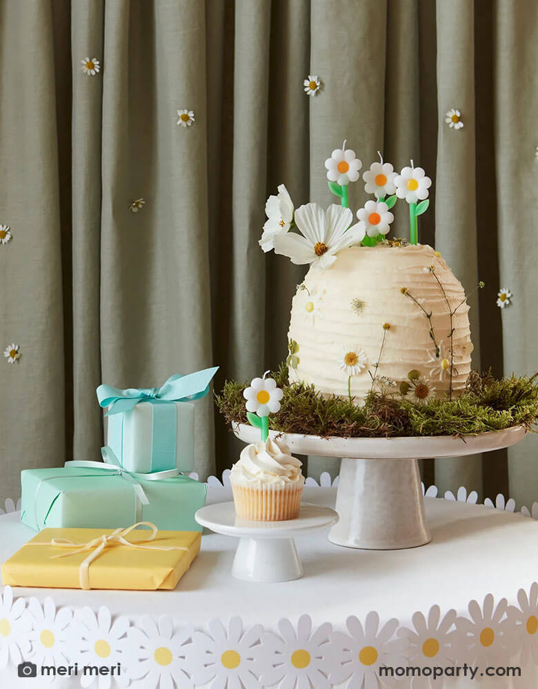 A wild daisy themed birthday party table featuring a buttercream cake decorated with real daisies on the side and topped with Momo Party's daisy candles from Meri Meri. There's also a daisy paper decoration wrap around the table which has some wrapped gifts on it.  In the back is a curtain decorated with real daisies flowers, making this an adorable inspo for girl's daisy or garden themed birthday party.