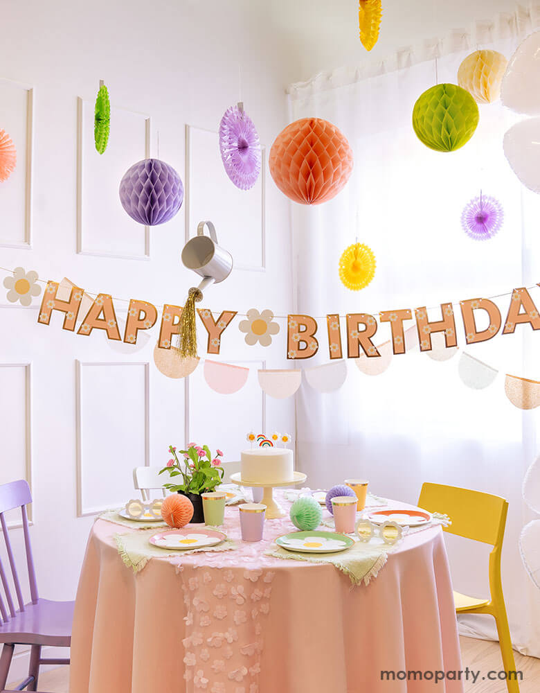 A bright and clean dining room decorated with pastel colored honeycombs and party fans hung from the ceiling. Momo Party's daisy Happy Birthday banner is hung underneath. On the table there are a various daisy themed tableware including plates, cups and birthday candles form Momo Party. Making this a dreamy, whimsical party set up for girl's birthday.