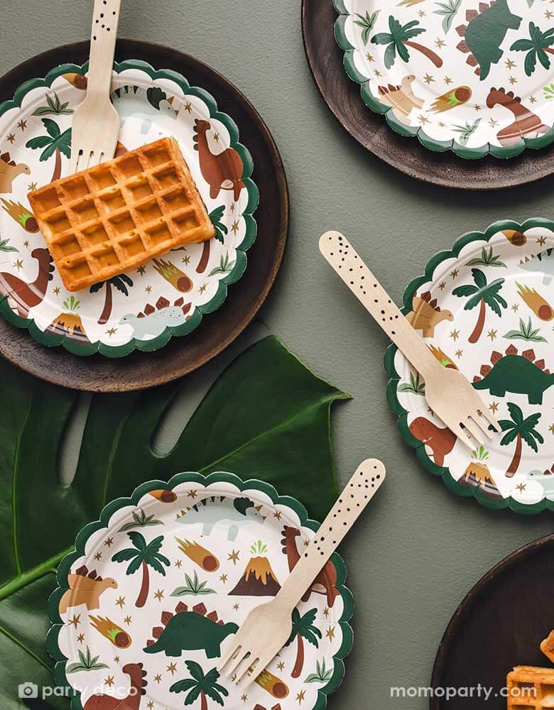 A dinosaur party table featuring Momo Party's 7" scalloped dinosaur side plates  in olive green background and palm leaves  around. With waffle snacks and wooden knives, these adorable dinosaur party plates are perfect for kid's dinosaur themed birthday celebrations.