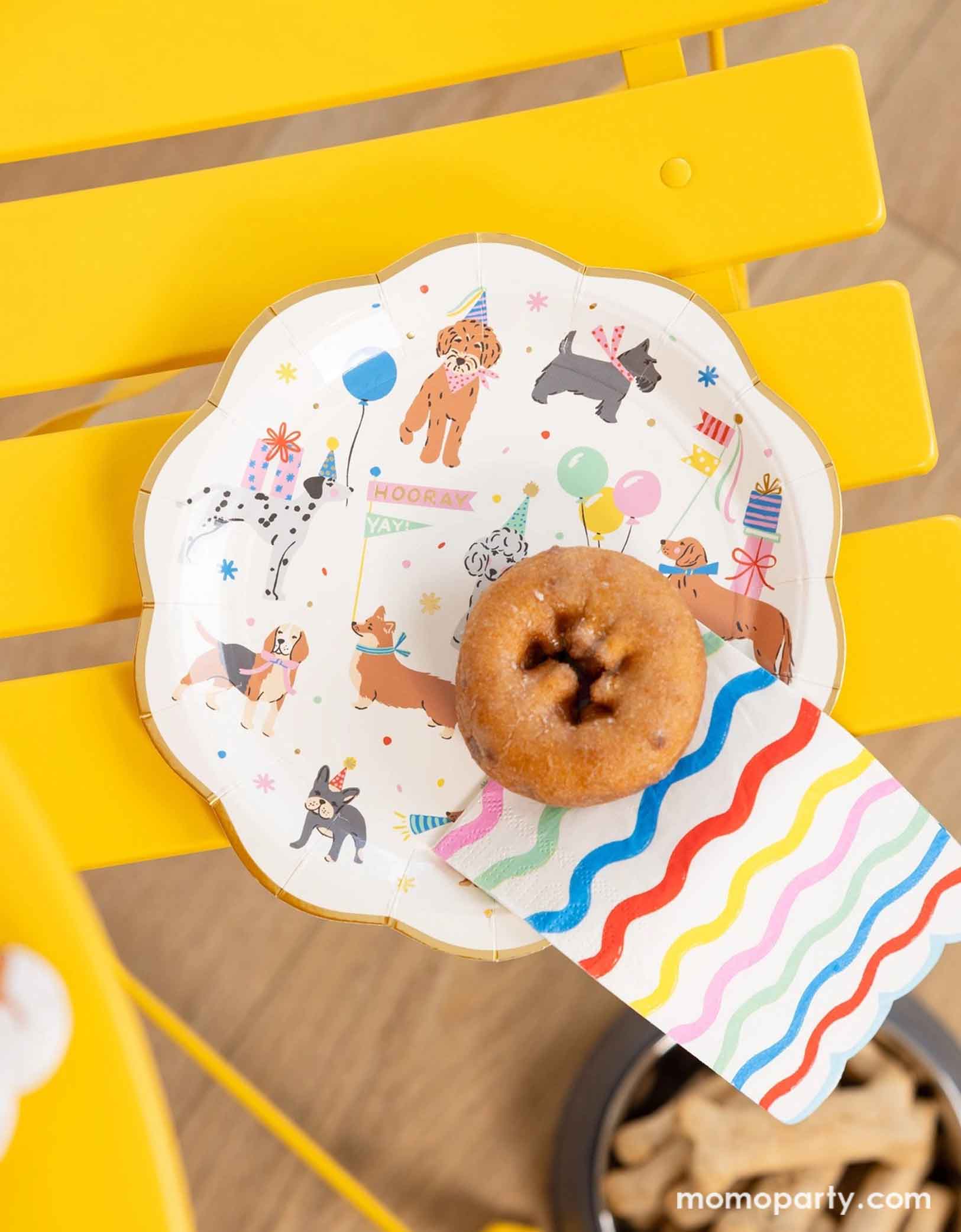 Momo Party's dog party plate featuring adorable illustrations of different breeds of dogs with party hats, balloons and birthday cakes on a bright yellow chair. On the plate there's a donut with ric rac rainbow colored napkin. Under the chair you can see a dog bowl filled with bone shaped treats for dogs.