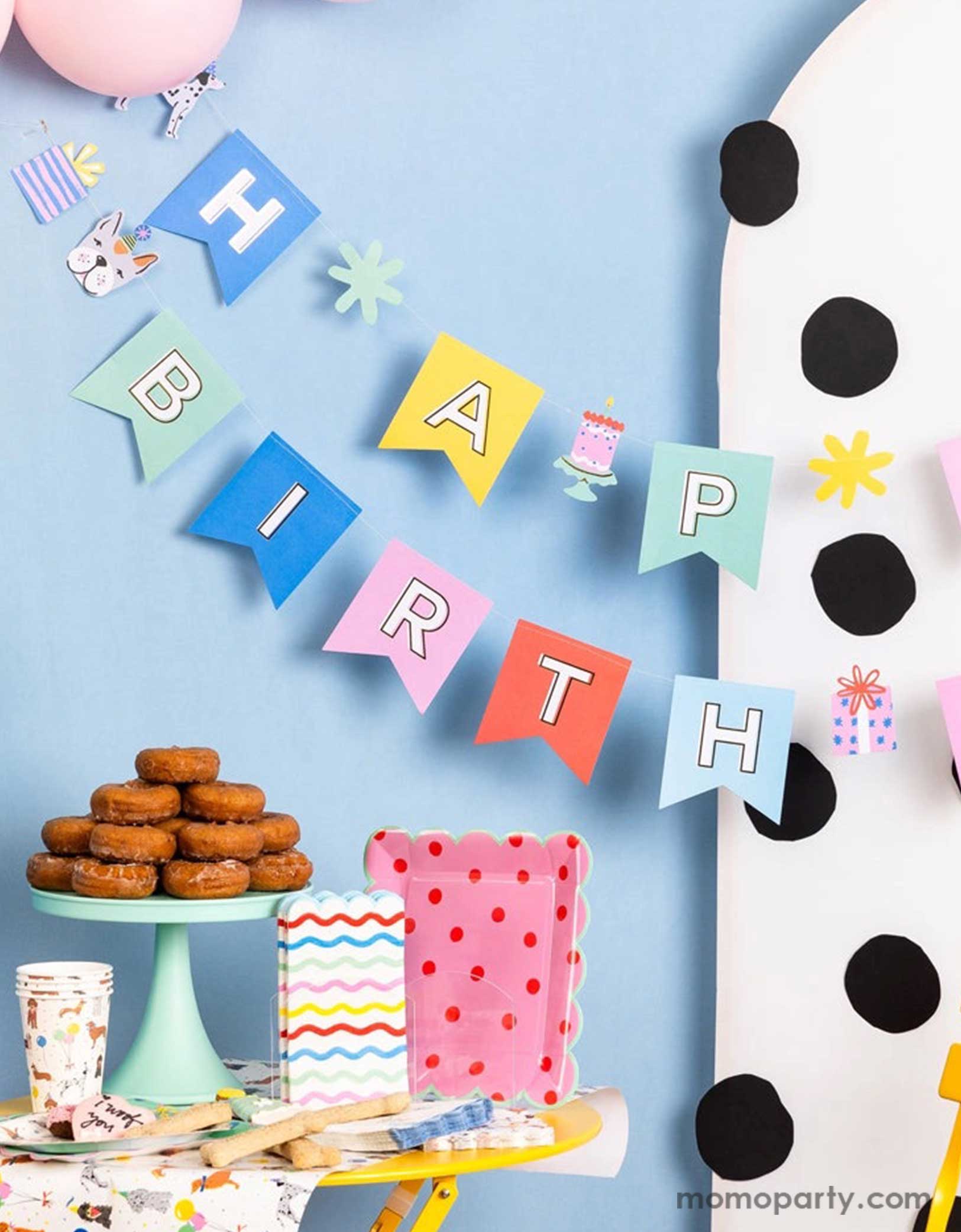 A corner of a festive and cheerful dog themed party featuring Momo Party's dog party tableware collection including dog themed party tablewares, the dog party Happy Birthday banner hung over a polka dotted party backdrop and party balloons. On the table there are some sugar cookies, treats and a donut tower,  ready for a fun and playful birthday celebration for a pet lover!